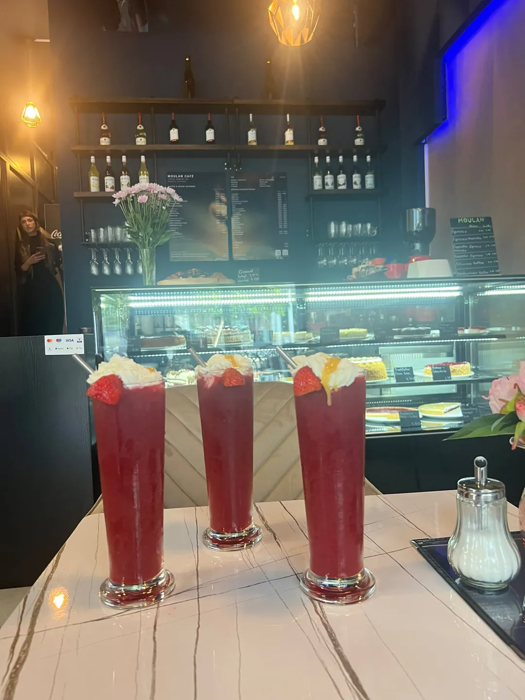 Drei Erdbeer-Drinks mit Sahnehaube auf einem Marmortisch vor der Kuchenvitrine im Moulan Café